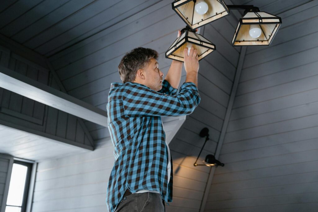 A man in a plaid shirt changes a light bulb on a ceiling lamp in a stylish, modern interior.