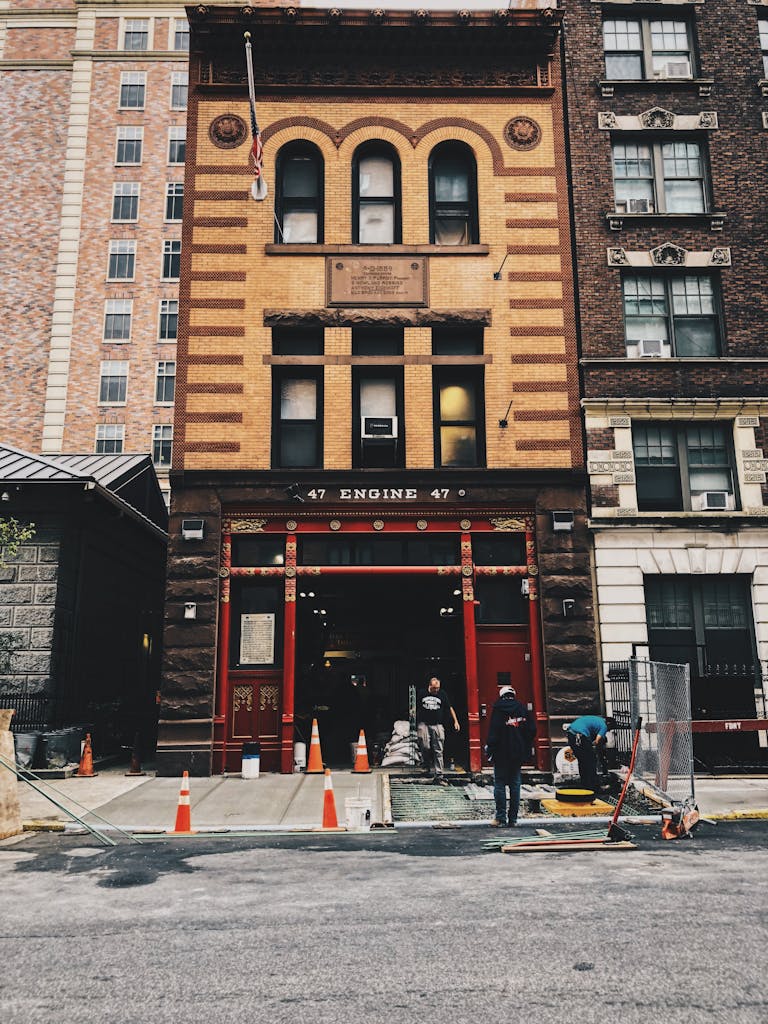 Classic fire station in urban setting with workers and construction cones on street.